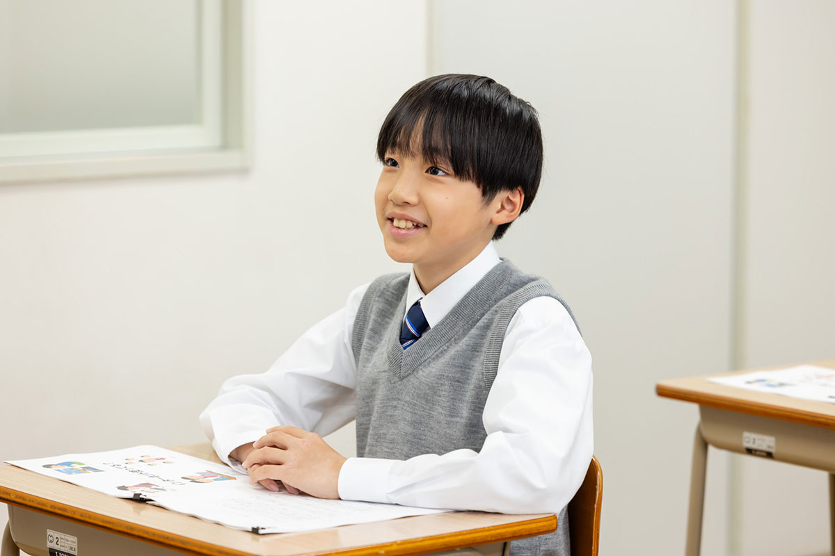 教室で教師が黒板の前で手を挙げて授業をしている様子です。生徒たちは机に座って先生の話を聞いています