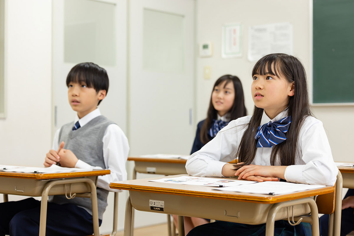 教師が生徒に向かって話しかけている場面です。教師は机に手をつき、親しみやすい態度で説明しています