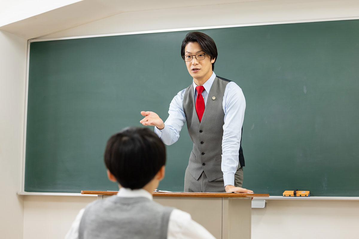 教室で教師が黒板の前で授業をしている様子です。生徒たちは机に座って先生の話を聞いています