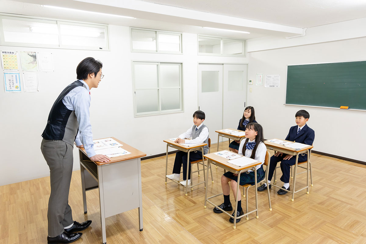教室で教師が黒板の前で授業をしている様子です。生徒たちは机に座って先生の話を聞いています