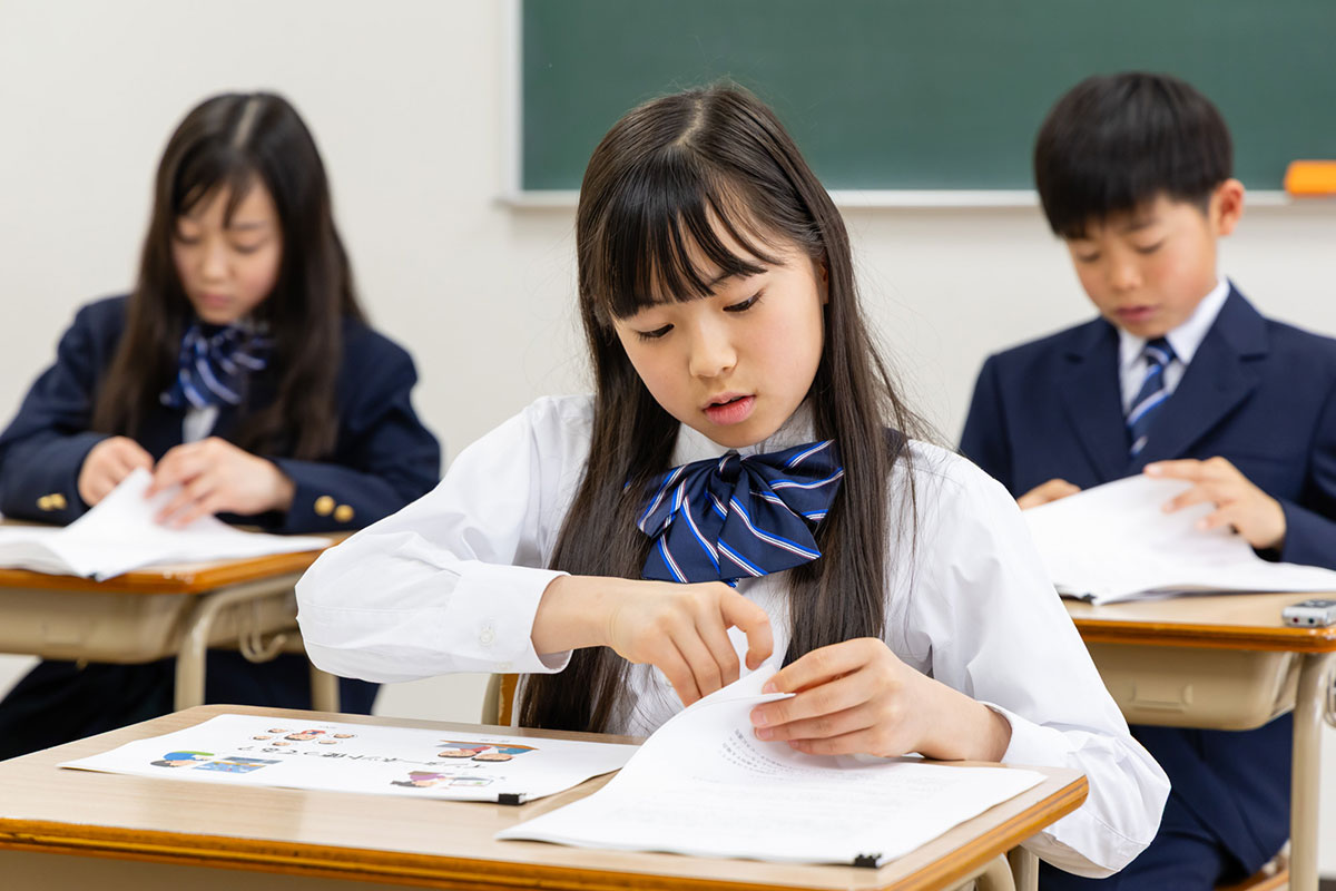 生徒たちがプリントを見ながら授業を受けている様子です。女の子が真剣にページをめくっています
