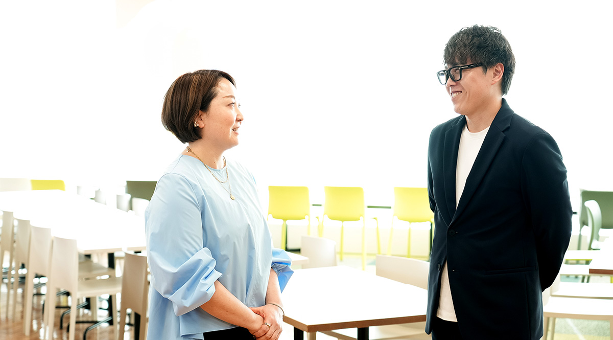 明るいオフィスで、番場と寺澤が向かい合って微笑みながら会話しています。リラックスした雰囲気です。