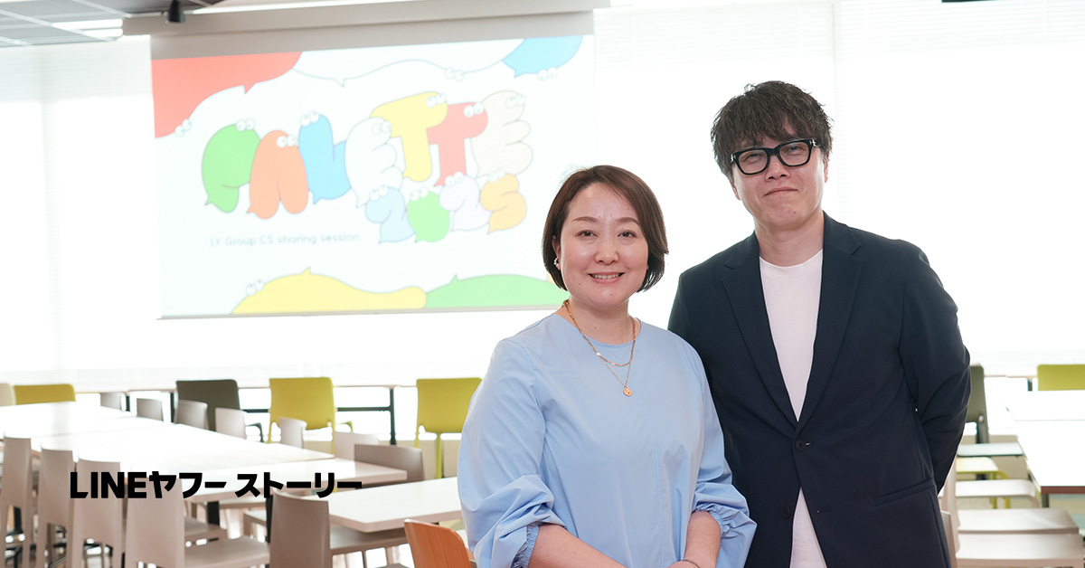 オフィスで、二人の人物がカメラに向かって微笑んでいます。背景には「PALETTE 2023」のスライドが表示されています。
