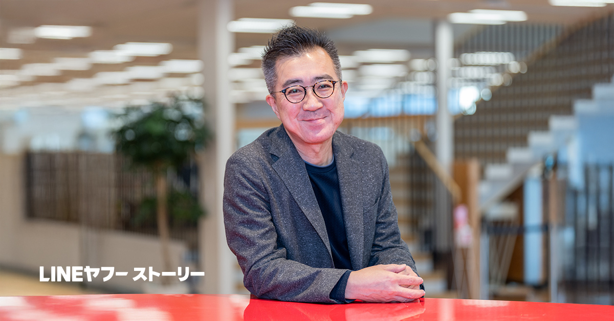 メガネをかけた兵頭CEOがオフィスのような場所で微笑んでいます。背景には階段と植物があります