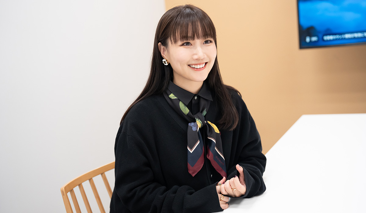 大塚愛さんのバストショット。黒いシャツとジャケットの上にカラフルな柄のスカーフを巻き、髪はストレートで前髪を下ろしている。明るい室内で白いテーブルに手を置いて座っており、微笑んでいる。