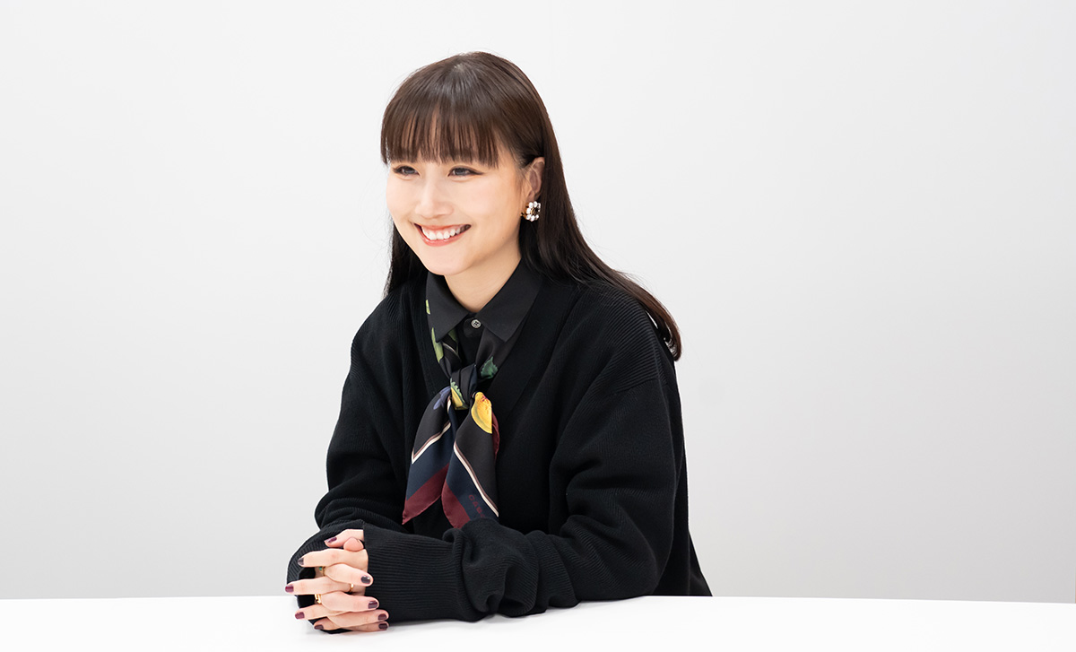 大塚愛さんのバストショット。手を組んで白いテーブルに置きながら、明るい笑顔を見せている。