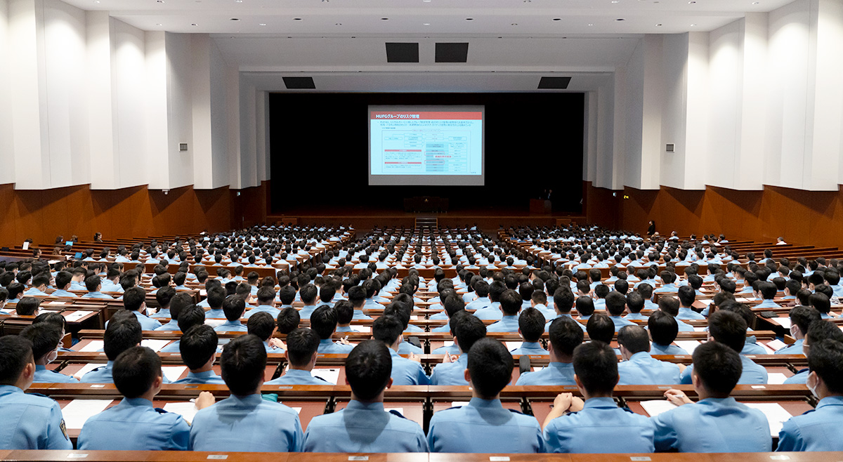 大講堂のような広い室内で、青い制服を着た多数の受講者が前方のスクリーンに向かって座っている様子。前方のスクリーンにはプレゼンテーション資料が投影され、壇上には発表者が立っている。