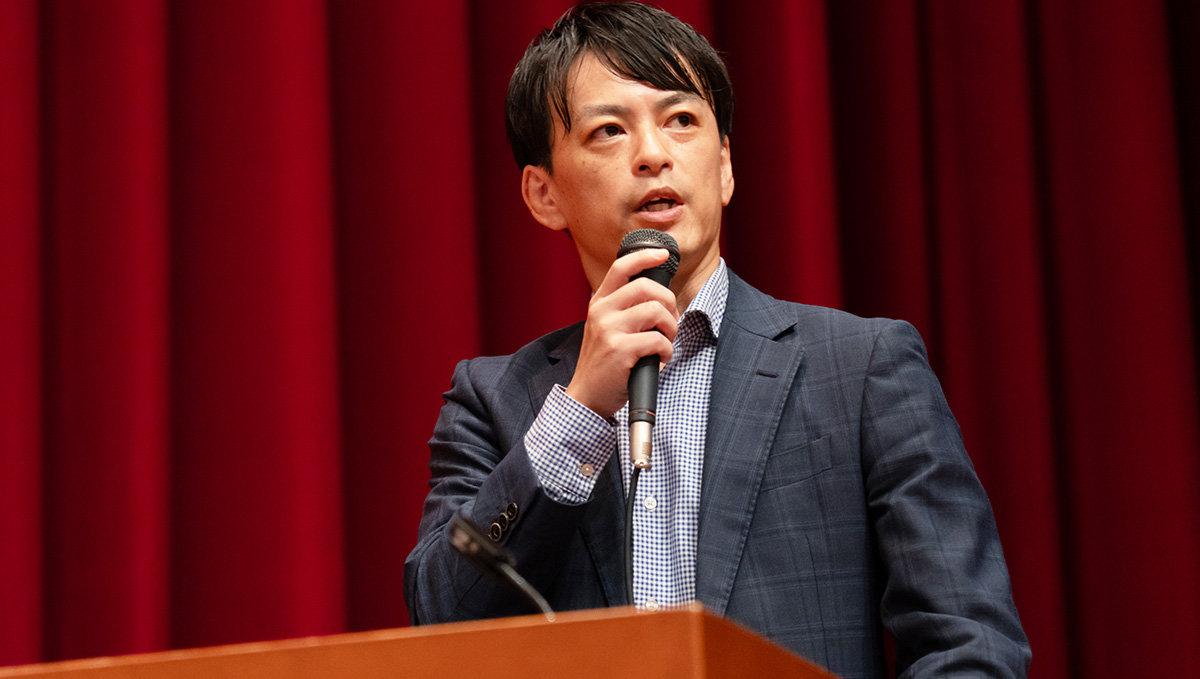 スーツを着た男性が壇上でマイクを持ち、発言している様子。背景には深い赤色のカーテンが広がり、スポットライトを浴びた男性がやや上を見ながら話している。