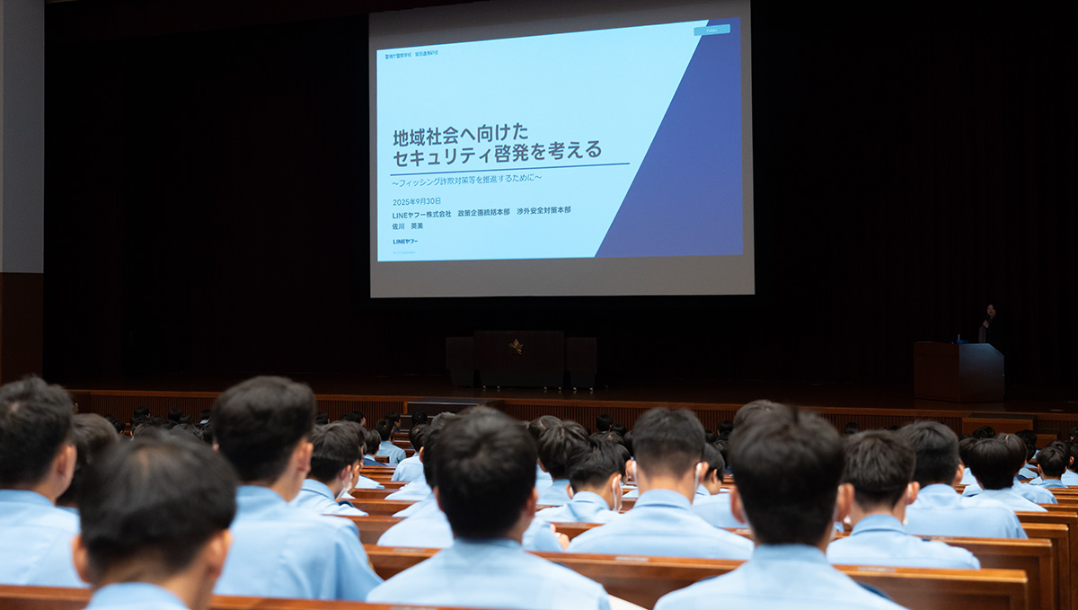 大講堂で青い制服を着た多数の受講者が着席し、前方のスクリーンを見ている様子。スクリーンには「地域社会へ向けたセキュリティ啓発を考える」と題したプレゼンテーションが映し出されており、壇上右手に登壇者が立っている。