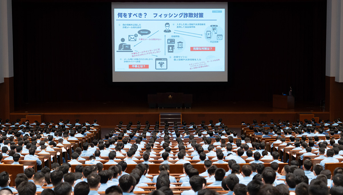 大講堂で青い制服を着た多くの受講者が前方のスクリーンに注目している様子。スクリーンには「何をすべき? フィッシング詐欺対策」と題したスライドが映し出され、詐欺メールの仕組みや対策の流れがイラスト付きで説明されている。壇上右側には講師が立ち、マイクを使って解説している。