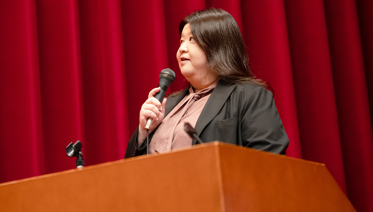 赤いカーテンの前で、女性が壇上に立ちマイクを手に話している様子。肩までの髪を下ろし、ブラウスと黒いジャケットを着ている。視線はやや斜め上を向いており、落ち着いた表情で講演をしている。