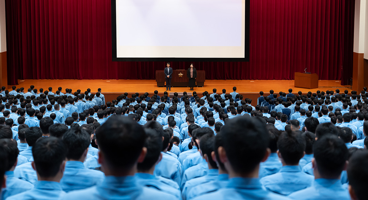 : 大講堂で青い制服を着た多数の人々が前方の舞台に注目している様子。舞台上にはスーツ姿の男性と女性が並んで立ち、背後には赤いカーテンと大型スクリーンがある。受講者たちは整列して起立している。