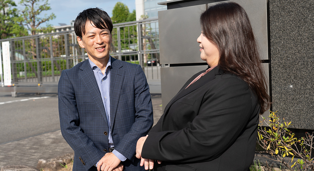 スーツ姿の男性と女性が屋外で向かい合い、笑顔で会話をしている様子。男性は青いチェック柄のジャケットを着て手を前で組み、女性は黒いジャケットを着て両手を軽く重ねている。