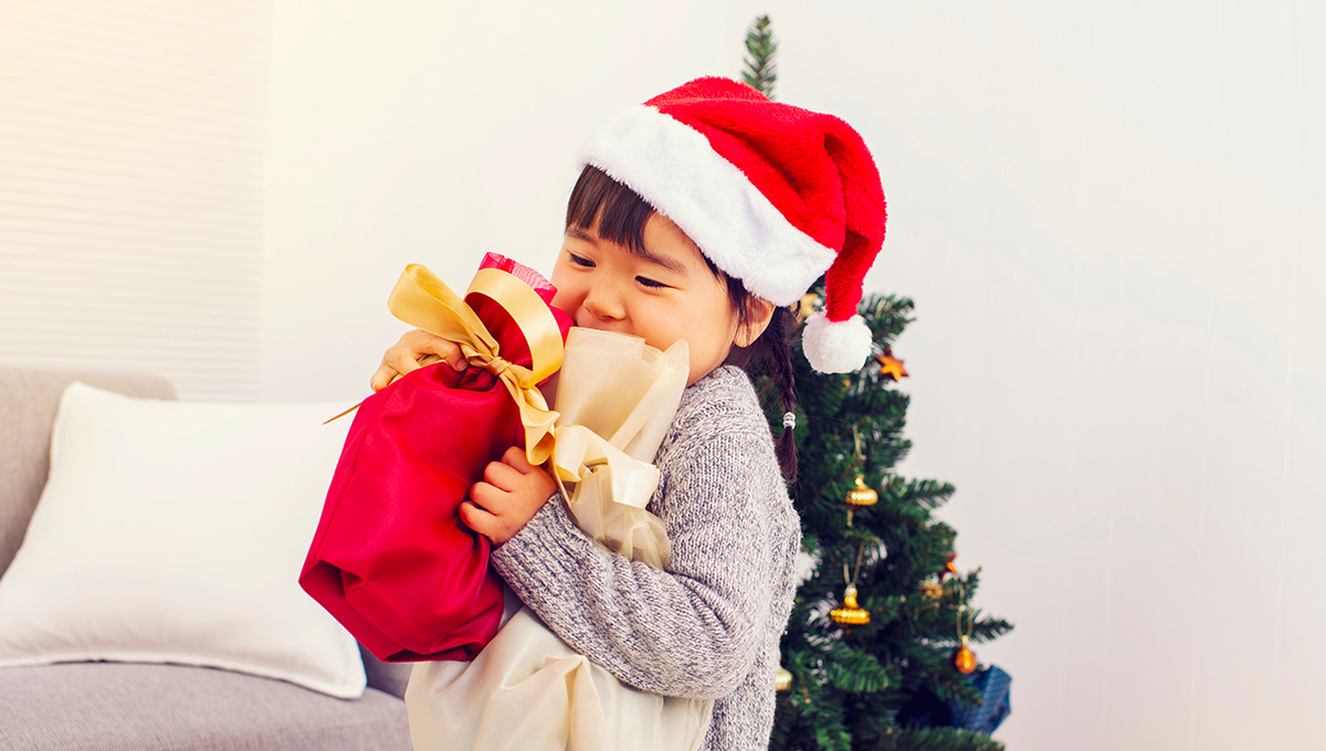 サンタ帽をかぶった子どもが、赤とベージュのプレゼント袋を胸に抱きしめて笑っている。子どもは灰色のセーターを着ており、背景には飾り付きのクリスマスツリーとソファが見える