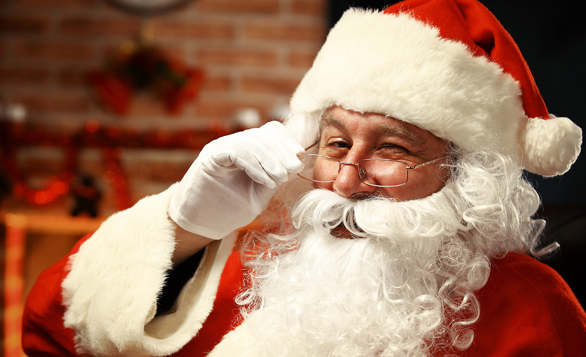 サンタクロースの衣装を着た人物のバストショット。赤い帽子と白いひげ、ふわふわした白い縁取りの赤いコートを着ており、右手に白い手袋をして眼鏡を持ち上げながらウインクしている。