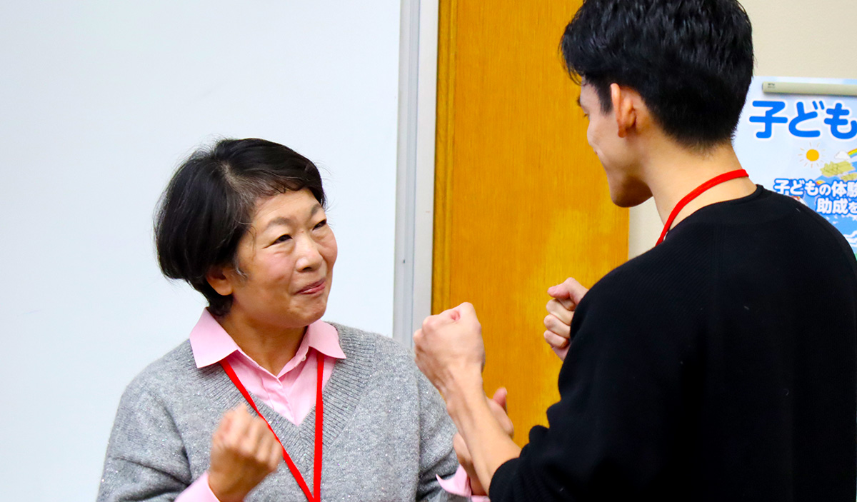 女性と男性が向かい合って話している様子。女性は灰色のニットにピンクのシャツを重ね着し、男性は黒い服を着ている。2人とも軽く両手を握って笑顔で会話している。