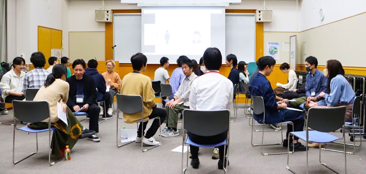 教室のような部屋で、複数の男女がペアになって椅子に座り、向かい合って会話している様子。