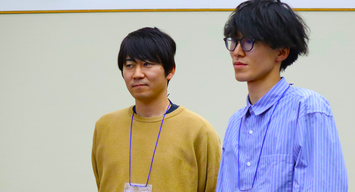 室内で並んで立つ2人の男性。左の人物は黄土色のセーターを着ており、右の人物は青と白のストライプ柄のシャツを着ている