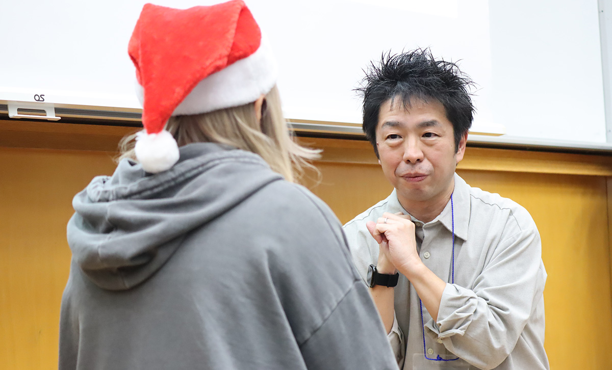 教室のような室内で、灰色のシャツを着た男性が前の人物と向かい合って話している。男性は両手を軽く握り、真剣な表情をしている。向かい合う人物はグレーのパーカーを着ており、赤いサンタ帽をかぶっている