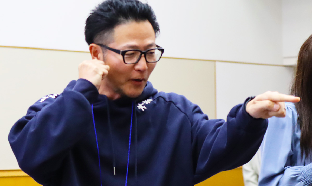 室内で男性が笑顔で指を差しながら、もう一方の手で拳を作って話している様子