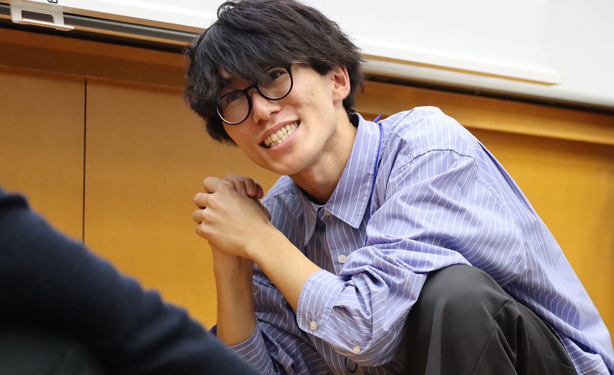 室内で、眼鏡をかけた男性がしゃがんで両手を組み、笑顔で相手を見つめている