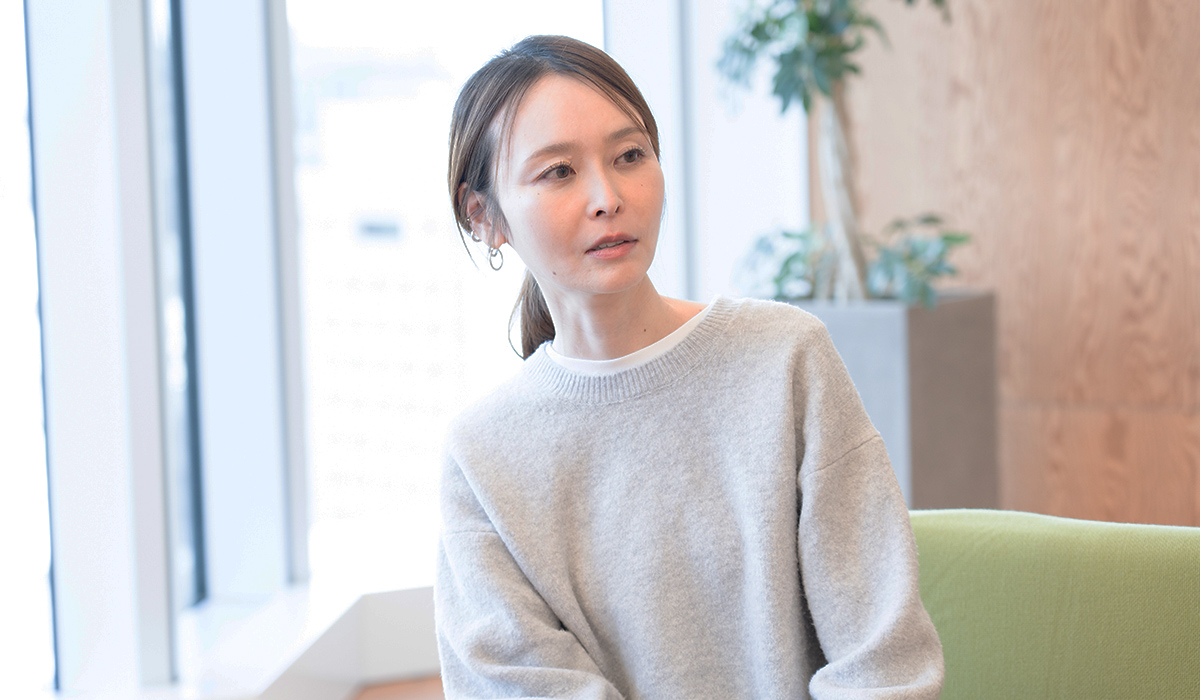 グレーのセーターを着た女性が、室内で横を向いて座っている様子です。背景には窓と観葉植物があります。