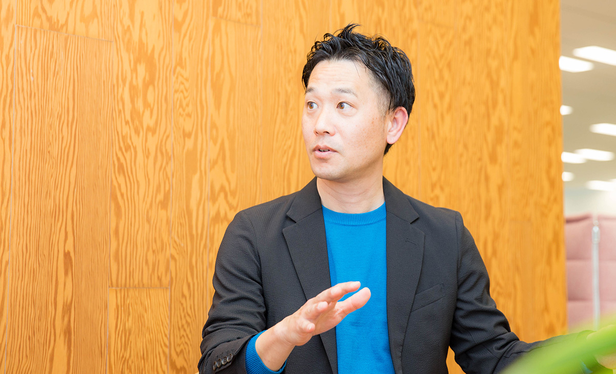 木目調の壁を背景に、小山が話している様子。視線を横に向けながら口を開き、右手を前に出して指を広げるような仕草をしており、説明や対話をしている。