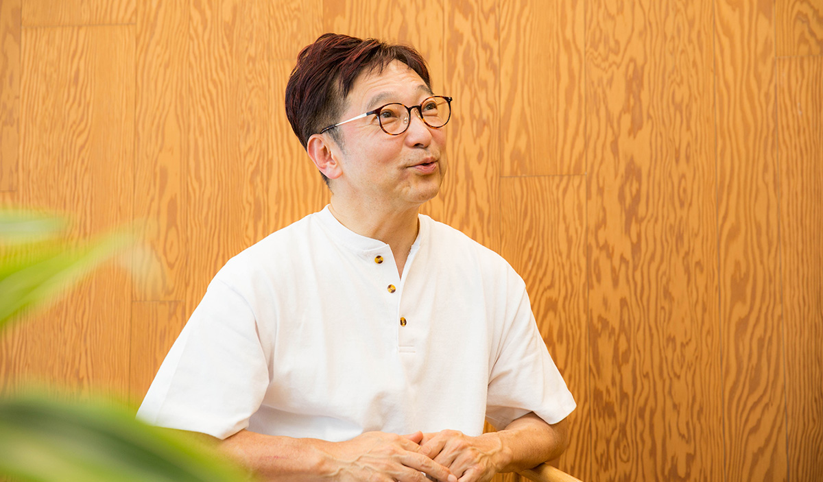 木目調の壁を背景に、斉藤さんが話している様子。斉やや上を見上げるように視線を向けながら口を開き、穏やかな表情で話している。両手は胸の前で軽く組まれ、リラックスした姿勢で椅子に腰掛けている。