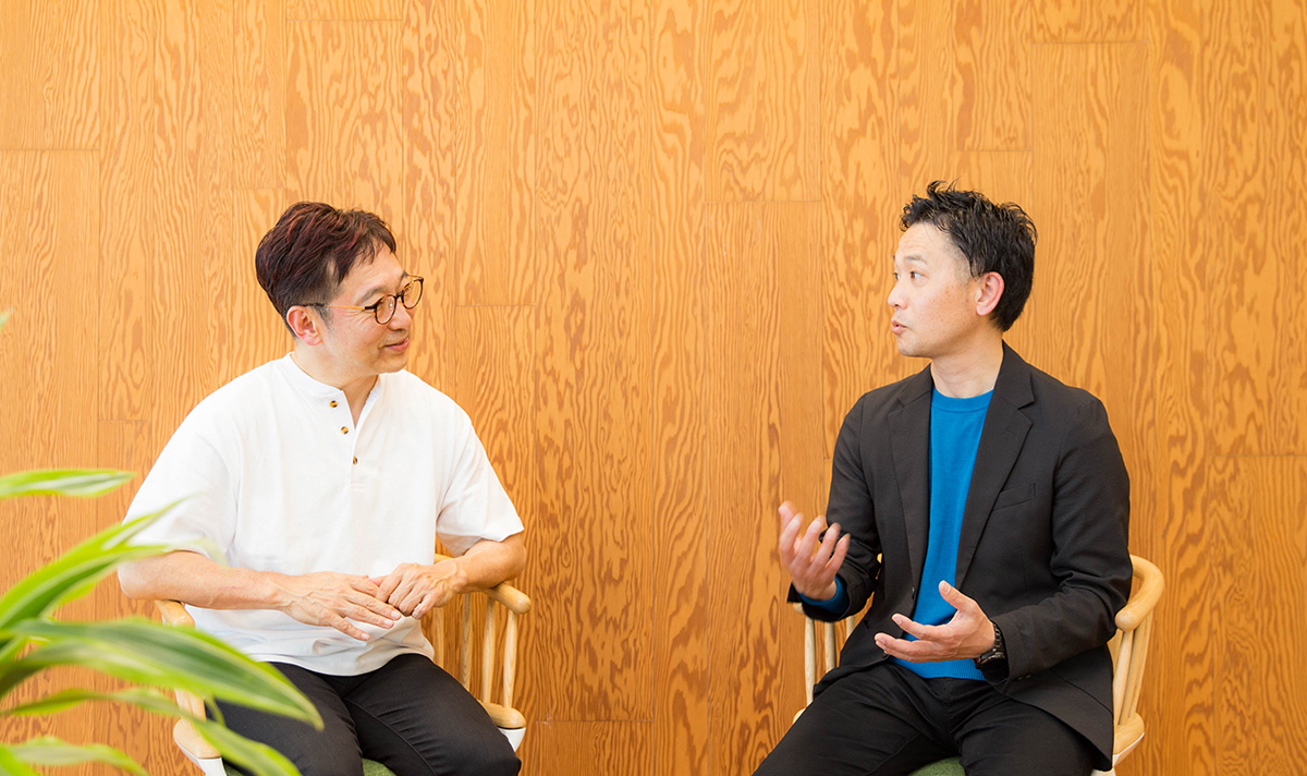 木目調の壁を背景に、斉藤さんと小山さんが向かい合って対話している。椅子に腰掛け、落ち着いた室内空間で真剣に意見を交わしている様子がうかがえる。