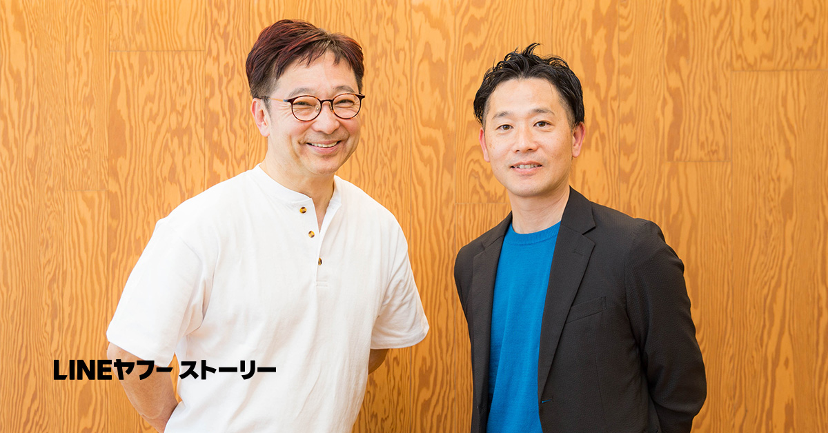 斉藤さんと小山のツーショット写真。左に立つ斉藤さんは、白い半袖シャツを着用し、丸眼鏡をかけて微笑んでいる。短く整えた髪で、両手を後ろに回した姿勢をしている。右に立つ小山は、黒いジャケットの下に青いTシャツを着用し、穏やかな表情で正面を向いている。背景は木目調の壁で、左下には「LINEヤフーストーリー」と黒い文字が配置されている。