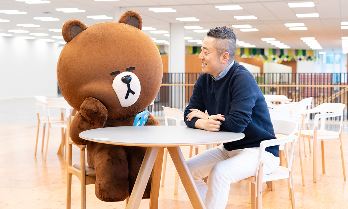 LINEキャラクターのブラウンと男性がオフィスのテーブルで向かい合って座っています。明るい空間で、リラックスした雰囲気が感じられます。
