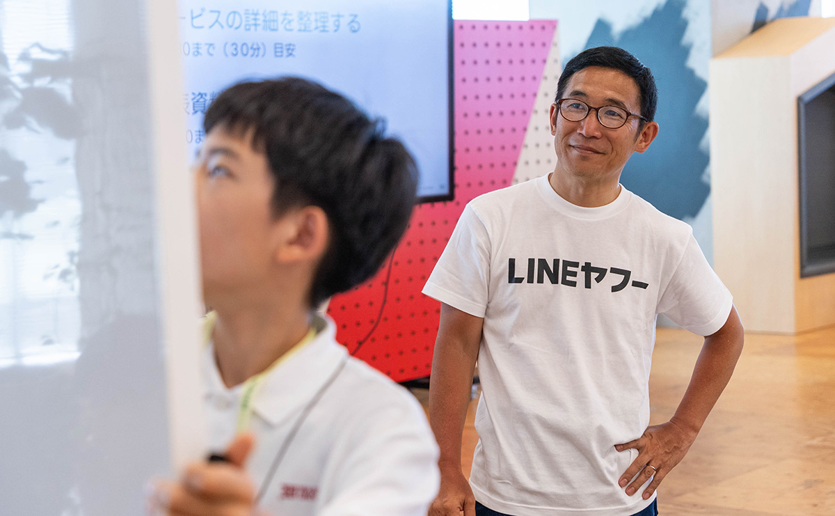白いTシャツを着た男性が、ホワイトボードに向かう少年を見守っています。背景にはプレゼンテーションが表示されています。