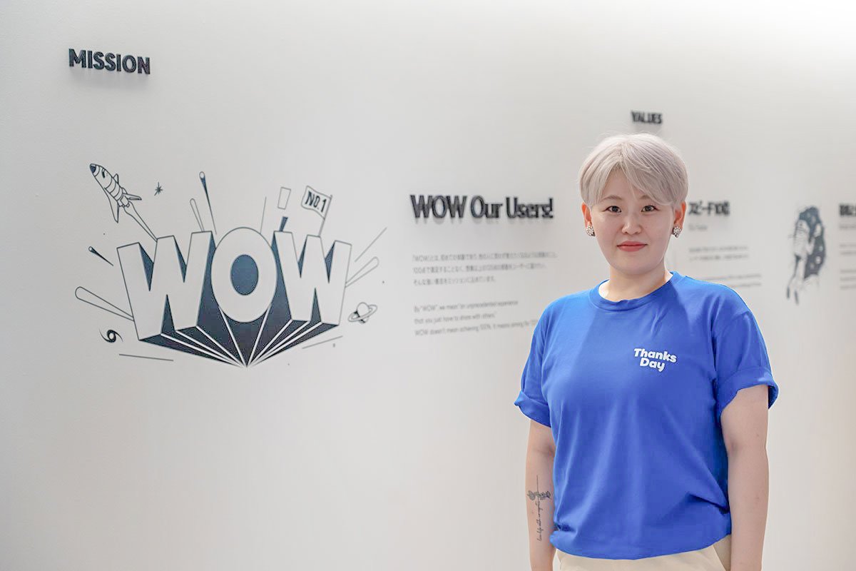 Thanks DayのスタッフTシャツを着た社員が、赤坂オフィスの壁画の前に立っている写真