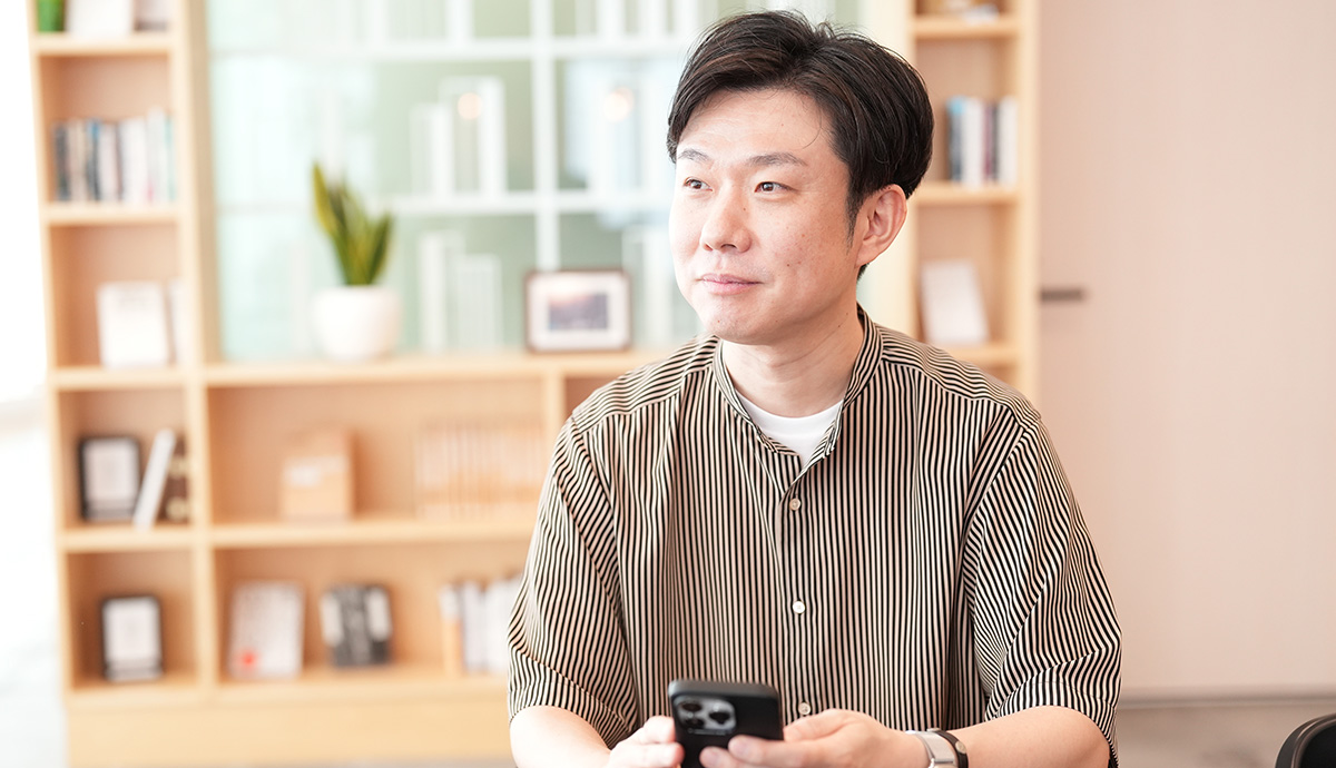 室内で携帯電話を手に持ち、微笑む男性。背景には本棚があり、リラックスした雰囲気です。
