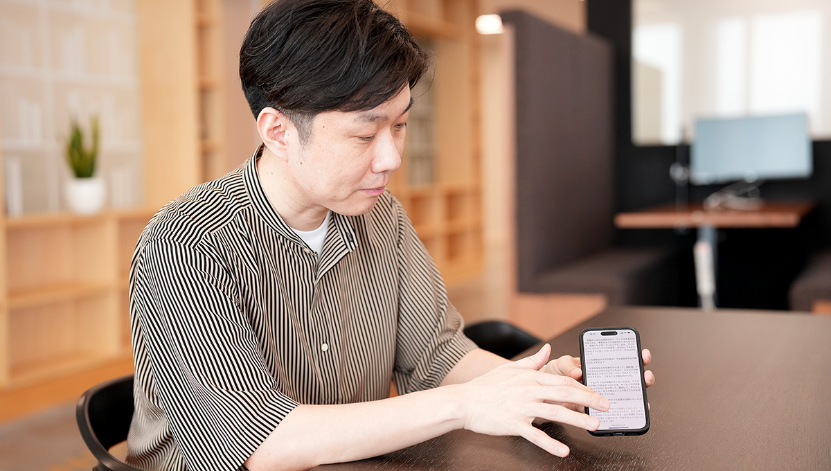室内でスマートフォンを操作する男性。机に座り、集中して画面を見ています。背景には本棚があり、落ち着いた雰囲気です。