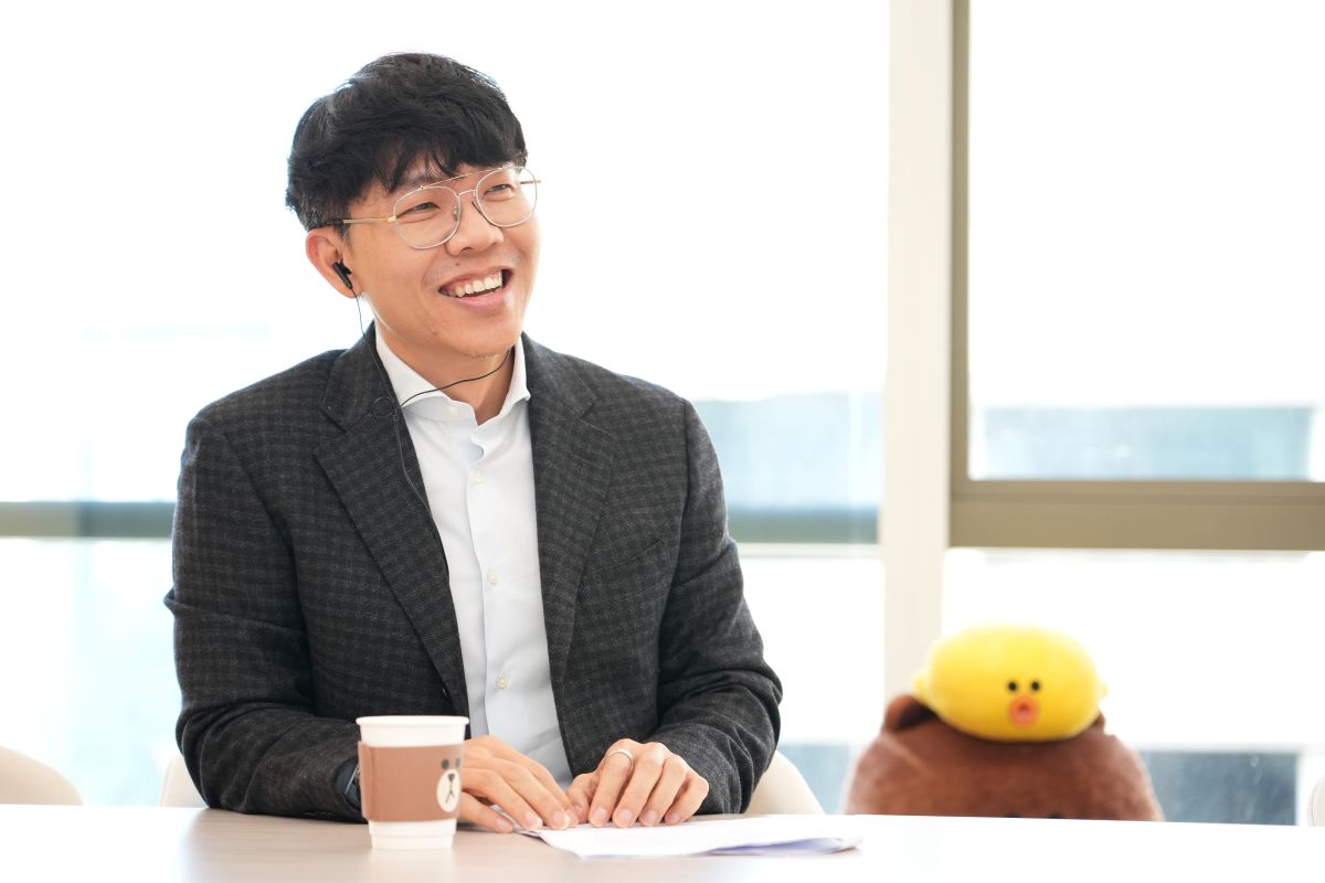 ネイトさんが笑顔で話している様子。LINEキャラクターの人形とカップがある。