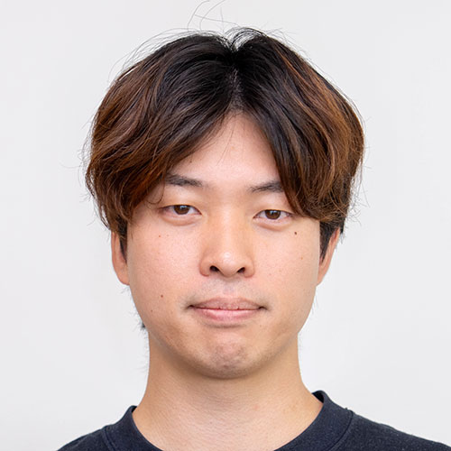 グレー背景の前で正面を向く豊田のバストショット。茶色がかった短髪を分けて整え、黒のトップスを着用し、口元を引き締めた真剣な表情をしている