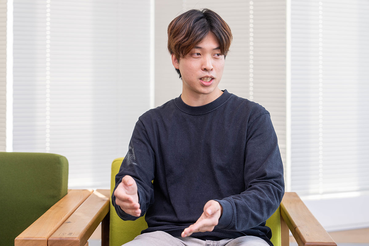 ブラインドを背景に椅子へ座る豊田の写真。茶色がかった短髪を分け、黒の長袖トップスを着用し、両手を前に差し出しながら説明するように話している。落ち着いた室内空間である