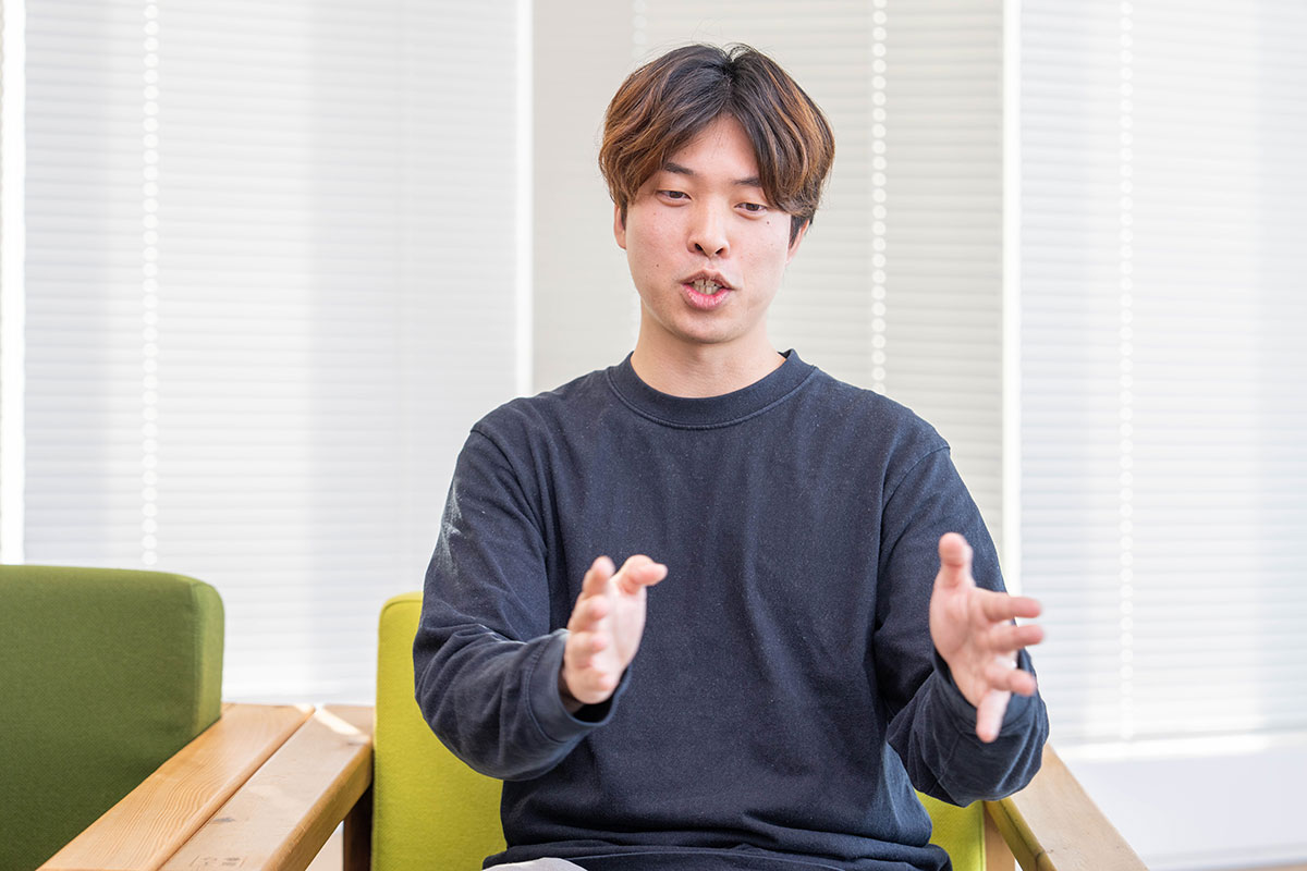 椅子に座る豊田が両手の幅を広げながら話す写真。黒の長袖トップスに淡い色のパンツを合わせ、真剣な表情で前方を見ている。背後には白いブラインドが並んでいる