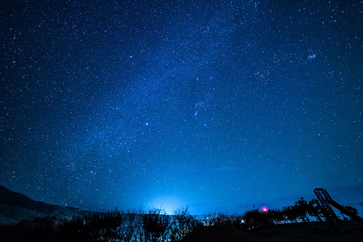 満天の星空が広がり、天の川の帯が流れる。地上のシルエットがわずかに浮かび、深い青が幻想的。
