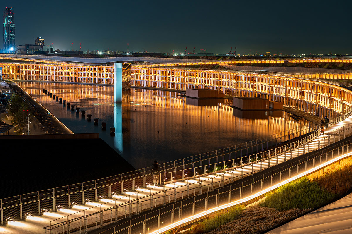 夜景の中、巨大な円形建築が金色に輝き水面に反射している。遠景に高層ビル群も見え、幻想的な雰囲気。