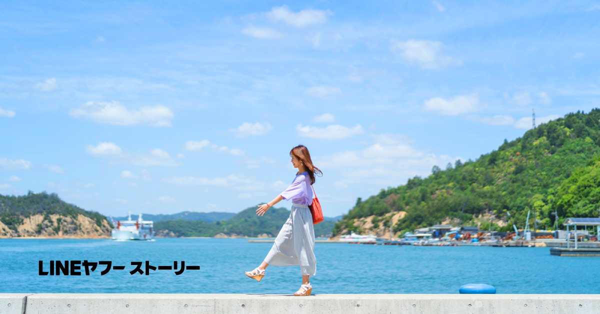 海辺の堤防を女性が軽やかに歩いている。青空と海が広がり、遠くに船と緑の島が見える爽やかな光景だ。