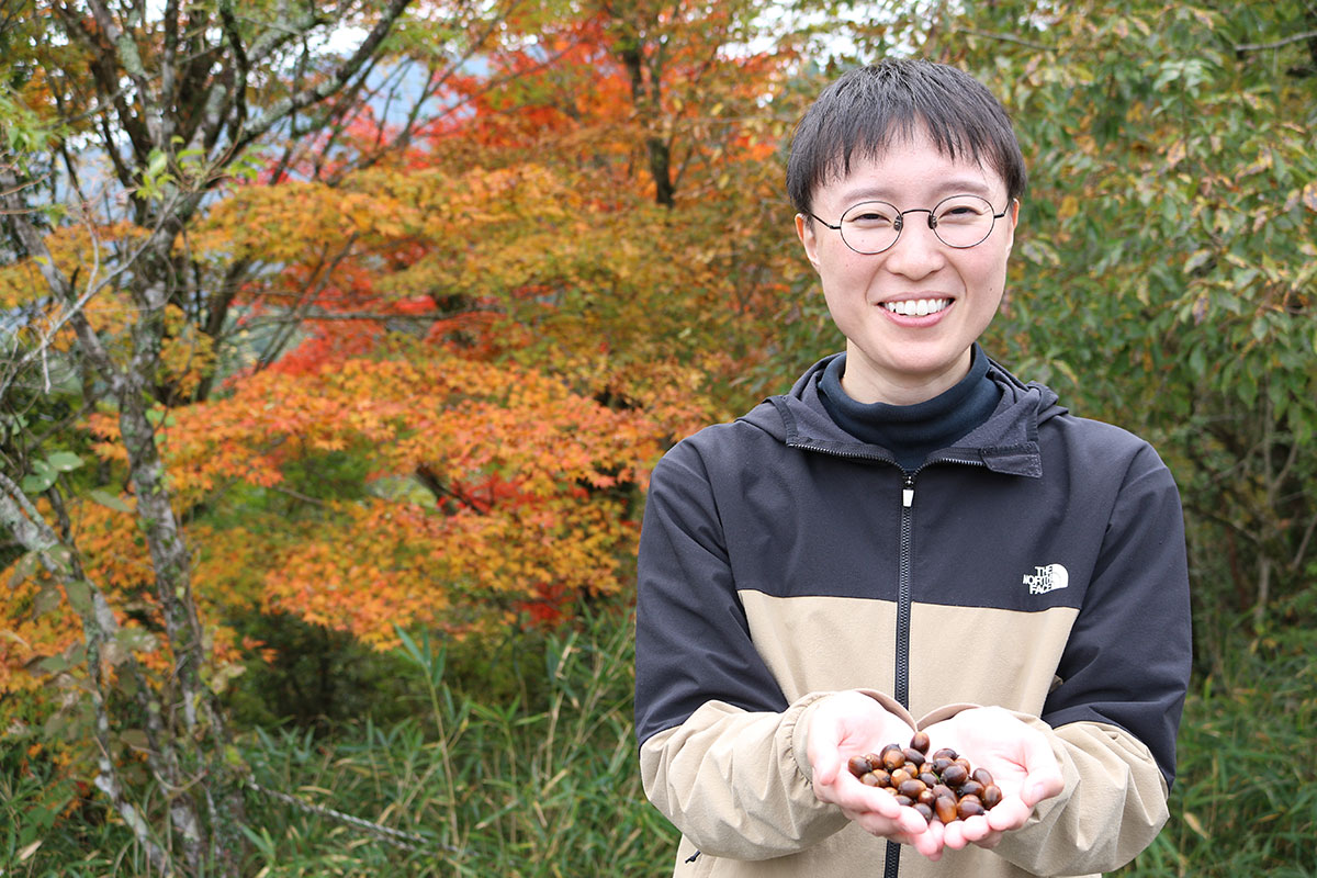 秋色の木々を背景に、丸眼鏡をかけた人物が笑顔で立っている。ベージュと黒の上着を着て、両手でどんぐりを差し出すように持っている。紅葉の鮮やかさが人物を引き立てている。