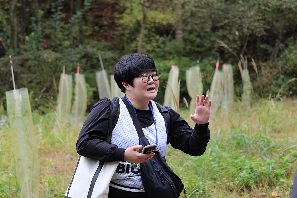 屋外の草地で、メガネをかけた人物が説明するように片手を上げて話している。後ろには植栽保護用の筒が並び、森の緑が広がっている。胸の前にはスマートフォンを持っている。