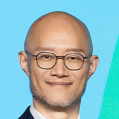 眼鏡をかけた男性のポートレート写真です。背景には地図が描かれており、ビジネスシーンを連想させます。