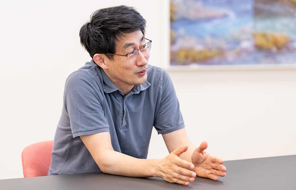 慎さんがテーブルに手を置き、熱心に話をしている様子です。背景には風景画が飾られています。