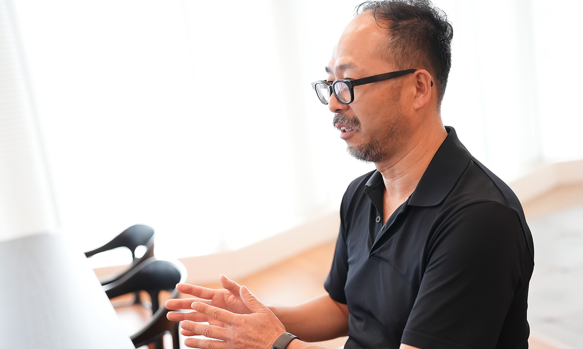黒い服を着た太田が、室内で熱心に話している様子です。背景には椅子が見え、明るい日差しが差し込んでいます。