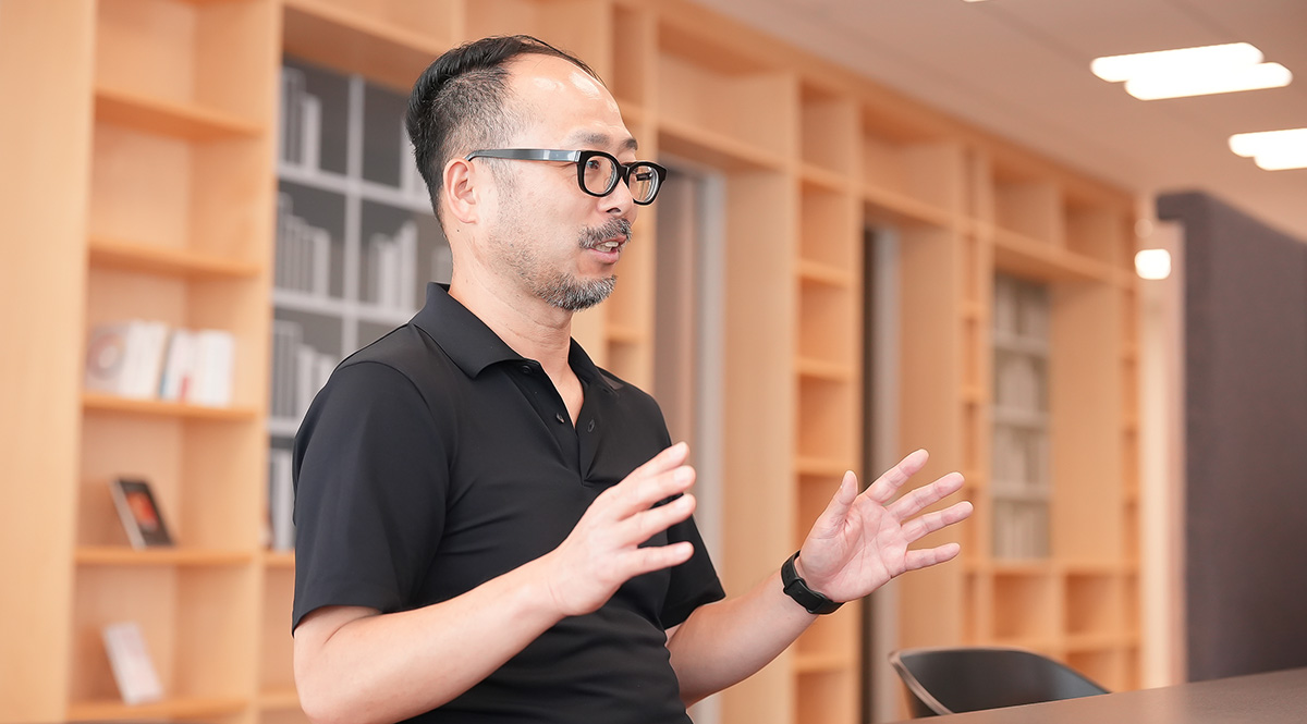 黒い服を着た太田が、室内で話をしている様子です。背景には本棚があり、明るいオフィスの雰囲気が感じられます。