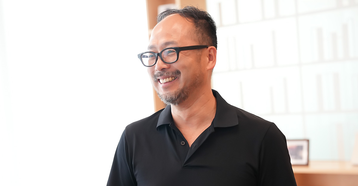 黒い服を着た太田が笑顔で立っている様子です。背景には本棚があり、明るい光が差し込んでいます。リラックスした雰囲気です。