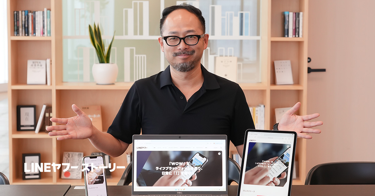黒い服を着た男性、太田が、スマートフォン、ラップトップ、タブレットを前にしてポーズを取っています。背景には本棚があります。